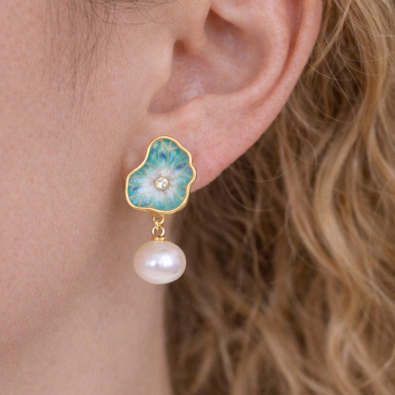May include: Gold earrings with a floral design, turquoise and white enamel, and a small diamond. Each earring has a dangling white pearl. The earrings are displayed on a person's ear.