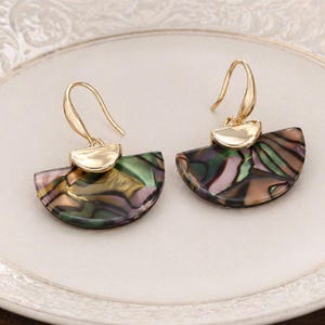 May include: A pair of gold-coloured earrings featuring a fan-shaped, iridescent, abalone-style design. The earrings have a gold hook and a small, half-moon shaped gold accent. The earrings are displayed on a white plate.