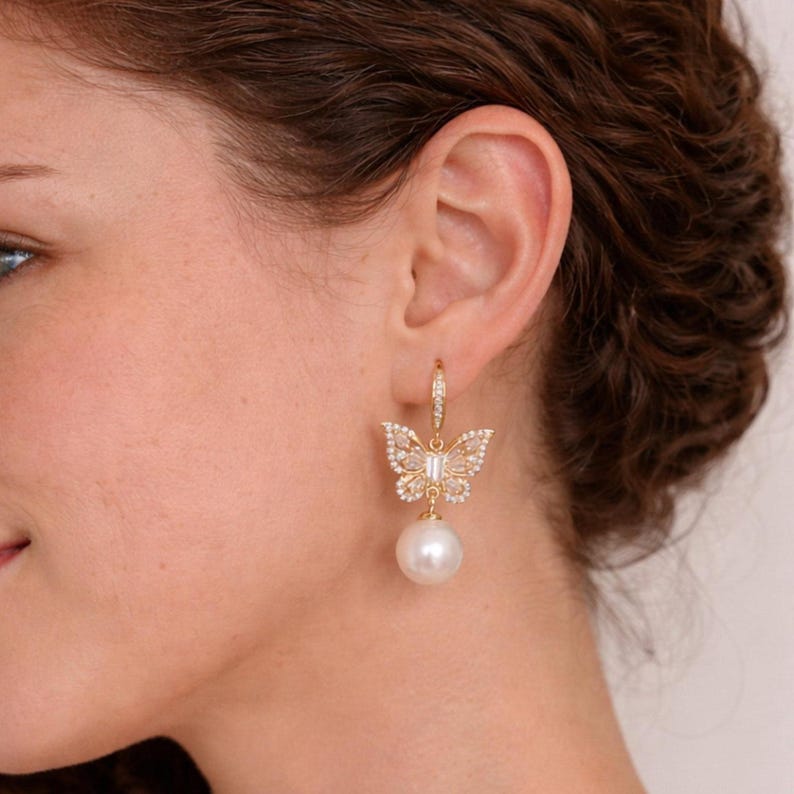 Pu&ograve; includere: Orecchini a farfalla dorati con una perla bianca pendente. Il design a farfalla &egrave; incastonato con piccole pietre preziose trasparenti. Gli orecchini sono attaccati a un piccolo cerchio con pietre preziose aggiuntive.