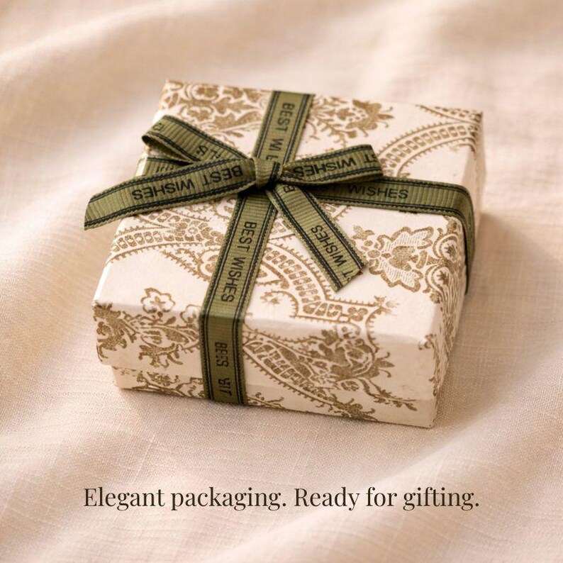 Pu&ograve; includere: Una piccola scatola regalo quadrata con sfondo bianco e motivo paisley dorato. La scatola &egrave; legata con un nastro verde con la scritta "BEST WISHES". Il testo "Elegant packaging. Ready for gifting." &egrave; in basso.