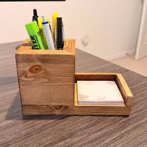 May include: A wooden desk organizer with a pen holder and a small notepad tray. The organizer is made of light-colored wood and contains pens, highlighters, and a notepad with floral accents. The design is simple and functional.