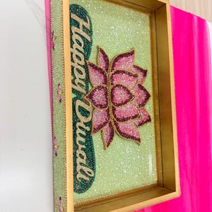 May include: A rectangular gold-toned tray featuring a light green glitter base. The tray displays a pink lotus flower design and the text "Happy Diwali" in gold script. The side of the tray is decorated with small pink floral accents.