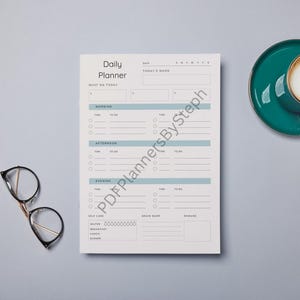 Op de afbeelding: Een dagplanner met een turquoise en wit ontwerp, met secties voor taken in de ochtend, middag en avond. Een turquoise koffiekopje en schotel, samen met een bril, liggen op een grijze ondergrond.