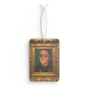 May include: Rectangular air freshener featuring a portrait of a man in a gold ornate frame. The man has dark eye makeup and a stern expression. The background is green. A white string is attached for hanging.