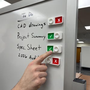May include: A white magnetic whiteboard with a To Do list featuring handwritten text: CAD drawings, Project Summary, Spec. Sheet, and 2026 Audit. Four rectangular toggle switches are attached to the board, each showing a green check or red X.