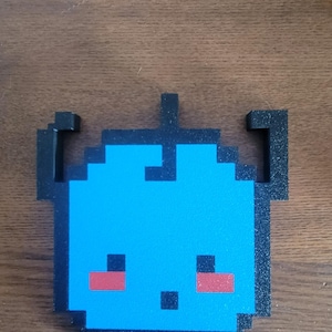 May include: A pixelated blue creature with black outlines and accents. The creature has red cheeks and a small black mouth. The design is reminiscent of retro video game characters. The object is likely a coaster or decorative item.