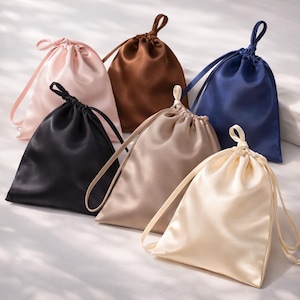 May include: Six satin drawstring bags in various colors: blush pink, brown, black, beige, navy blue, and cream. Each bag has a matching satin drawstring closure. The bags are arranged on a white surface.