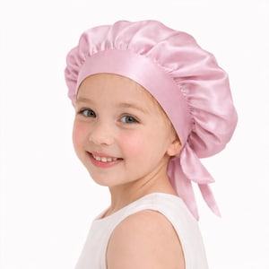 May include: A light pink satin bonnet with a wide band and a tie closure. The bonnet is designed to protect hair and is made of a shiny, smooth fabric. The bonnet is shown on a young child.