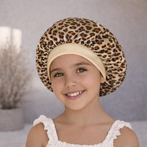 May include: A satin hair bonnet with a leopard print pattern. The bonnet features a tan elastic band and a rounded crown. The image shows a young person wearing the bonnet, smiling at the camera. The background is a soft, neutral color.