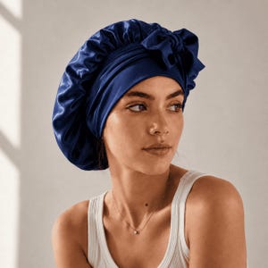 May include: A navy blue satin bonnet with a gathered crown and a bow detail. The bonnet is designed to protect hair and maintain hairstyles. The image shows the bonnet on a person, highlighting its shape and color.