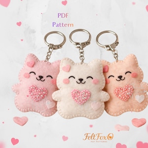Puede incluir: Tres llaveros de fieltro con forma de gato en rosa, blanco y melocotón, cada uno con un adorno en forma de corazón y una cara sonriente. Los llaveros están unidos a anillas plateadas. La imagen incluye el texto "PDF Pattern" y el logotipo de FeltFoxO.
