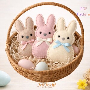 May include: A wicker basket contains three felt bunny rabbits in beige, pink, and cream, each with a bow. The basket also holds decorative eggs and straw. The image has the text "PDF Pattern".