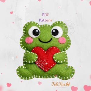Könnte beinhalten: Ein grüner Frosch aus Filz, der ein rotes Herz hält. Der Frosch hat große schwarze Augen, rosa Wangen und weiße Nähte. Das Bild enthält den Text "PDF Pattern" und das Felt Fox Logo.