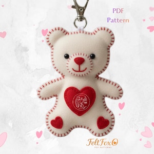 May include: A white felt teddy bear keychain with a red heart and accents. The bear has black eyes, a red nose and mouth, and red stitching. The heart on the bear's chest has a white logo. The keychain has a silver clasp. Text on the image says "PDF Pattern" and "Felt Fox".