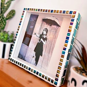Op de afbeelding: Een ingelijste kunstdruk met een zwart-wit afbeelding van een meisje met een paraplu in de regen. De witte lijst is versierd met kleurrijke mozaïektegels. Het kunstwerk wordt op een houten oppervlak getoond.