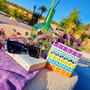 Può includere: Un cubo a mosaico colorato con motivi geometrici di quadrati, cerchi e triangoli nei toni del viola, blu, giallo e arancione. Occhiali da sole neri su un libro. Una bottiglia di vetro verde sullo sfondo.