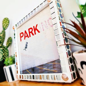 Op de afbeelding: Een ingelijst kunstwerk met een witte mozaïekrand. Het kunstwerk toont het woord "PARK" in het rood, met een klein figuurtje op een schommel. Het frame is versierd met kleine spiegels en kleurrijke accenten.