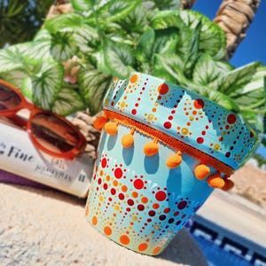 Może przedstawiać: Turkusowa doniczka pomalowana we wzory w pomarańczowe i czerwone kropki oraz pomarańczowe wykończenie z pomponami. Doniczka jest ozdobiona różnymi rozmiarami i kolorami kropek. Para pomarańczowych okularów przeciwsłonecznych i książka w tle.