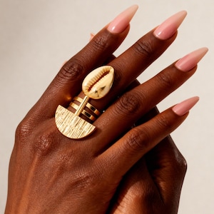 Puede incluir: Anillo dorado con diseño de concha y forma de media luna. El anillo tiene una superficie texturizada y se lleva en una mano con uñas largas y rosas. Una pieza de joyería llamativa.