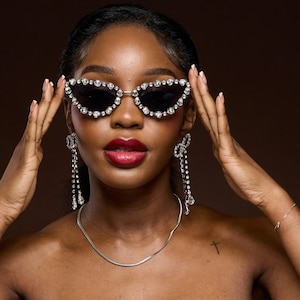 Op de afbeelding: Cat-eye zonnebril met een zilver montuur en zwarte glazen, versierd met sprankelende strass-steentjes. Het model draagt ook lange, bungelende oorbellen met kristallen accenten en een zilveren ketting. De achtergrond is effen bruin.