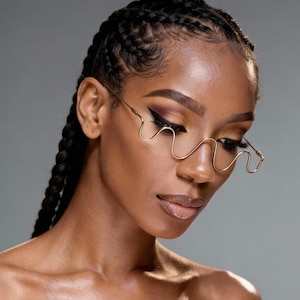 Könnte beinhalten: Eine Frau mit dunkler Haut und geflochtenem Haar trägt eine goldfarbene Brille mit wellenförmigem Rahmen. Die Brille hat ein einzigartiges, durchgehendes Design. Die Frau hat dunkles Augen-Make-up und blickt nach unten.