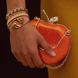 May include: An orange, apple-shaped clutch with a gold chain strap and stem. The clutch is covered in small, sparkling embellishments. A gold bracelet and two gold bangles are visible.