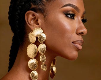 May include: A woman with dark skin and braided hair is wearing large, gold, hammered-metal earrings. The earrings are composed of multiple, overlapping circular shapes that hang down from the earlobes. The background is a warm, neutral color.