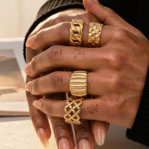 May include: A hand displaying four gold rings. The rings showcase diverse designs: a chain link, a geometric diamond pattern, horizontal ridges, and an openwork lattice. The rings are set against a neutral backdrop.