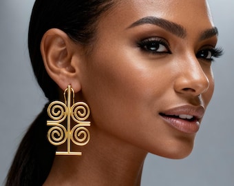 May include: A woman with dark hair and makeup is wearing large, gold-colored earrings. The earrings have a geometric design with a vertical bar and four spiral shapes. The background is a soft gray.
