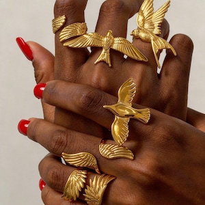 Pode incluir: Anéis dourados com motivos de pássaros e asas. Os anéis são exibidos em uma mão com esmalte de unhas vermelho.