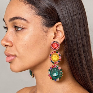 May include: Dangling earrings with a vibrant, multi-colored design. Each earring features three large, jewel-toned circular stones: red, yellow, and green, surrounded by smaller, colorful gems. The earrings have a gold-colored metal setting.