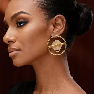 Op de afbeelding: Een vrouw draagt gouden oorringen met een spiraalontwerp en een horizontale balk. Ze draagt ook een fijne gouden ketting. De oorbellen zijn een opvallend sieraad, dat een vleugje elegantie aan haar look toevoegt.