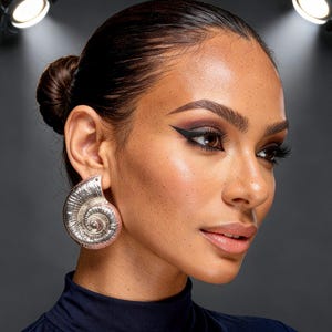 Könnte beinhalten: Eine Frau trägt große, silberne Ohrringe in Muschelform. Die Ohrringe sind mit Spiralmustern versehen. Sie trägt ein dunkelblaues Oberteil und hat dunkles Augen-Make-up. Der Hintergrund ist dunkelgrau.