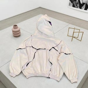 K&ouml;nnte beinhalten: Ein cremefarbener Kapuzenpullover mit schwarzen, rissartigen Linien auf der Vorderseite und den &Auml;rmeln. Die Kapuze hat einen lila Akzent. Eine rosa Vase und eine goldene geometrische Skulptur sind ebenfalls im Bild.