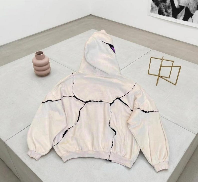 K&ouml;nnte beinhalten: Ein cremefarbener Kapuzenpullover mit schwarzen, rissartigen Linien auf der Vorderseite und den &Auml;rmeln. Die Kapuze hat einen lila Akzent. Eine rosa Vase und eine goldene geometrische Skulptur sind ebenfalls im Bild.