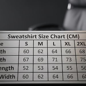 K&ouml;nnte beinhalten: Ein silbernes Metallschild mit einer Sweatshirt-Gr&ouml;&szlig;entabelle in Zentimetern. Die Tabelle enth&auml;lt die Gr&ouml;&szlig;en S bis 3XL mit Ma&szlig;en f&uuml;r Breite, L&auml;nge, &Auml;rmell&auml;nge und Schulterbreite. Das Schild liegt auf einer dunklen Oberfl&auml;che.