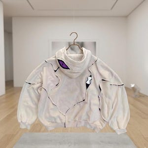 K&ouml;nnte beinhalten: Cremefarbener Kapuzenpullover mit einzigartigem Design, mit schwarzen Stepplinien und lila Augendetails auf der Kapuze. Der Hoodie hat eine lockere Passform und h&auml;ngt an einem Metallb&uuml;gel.