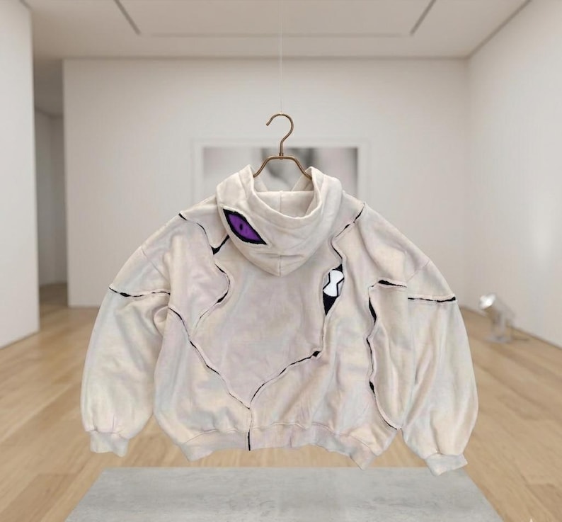 K&ouml;nnte beinhalten: Cremefarbener Kapuzenpullover mit einzigartigem Design, mit schwarzen Stepplinien und lila Augendetails auf der Kapuze. Der Hoodie hat eine lockere Passform und h&auml;ngt an einem Metallb&uuml;gel.