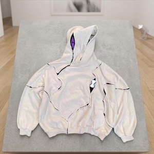 K&ouml;nnte beinhalten: Cremefarbener Kapuzenpullover mit schwarzen Akzentlinien und einem einzigartigen Design. Die Kapuze hat lila Akzente, und eine schwarz-wei&szlig;e Grafik befindet sich auf der Vorderseite. Der Pullover hat eine lockere Passform und lange &Auml;rmel.