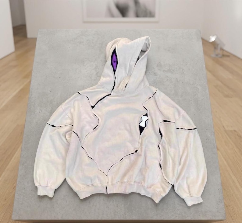 K&ouml;nnte beinhalten: Cremefarbener Kapuzenpullover mit schwarzen Akzentlinien und einem einzigartigen Design. Die Kapuze hat lila Akzente, und eine schwarz-wei&szlig;e Grafik befindet sich auf der Vorderseite. Der Pullover hat eine lockere Passform und lange &Auml;rmel.