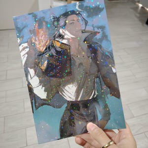 Puede incluir: Una impresión holográfica que presenta un personaje con cabello largo y oscuro y un atuendo oscuro. La obra de arte tiene un tema de fantasía con un fondo azul y verde azulado y acentos brillantes. La impresión es sostenida por una mano con un anillo de oro.