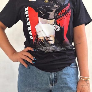 May include: A black short-sleeve t-shirt featuring a graphic print of an inverted face with a white blindfold, set against a red background. The word PARADISE is printed vertically in white, accompanied by small text lines in English and Korean.