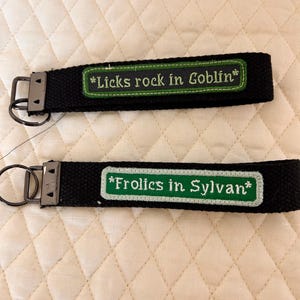 May include: Two black fabric wristlet keychains with dark metal hardware. The top keychain features a green embroidered patch with the text *Licks rock in Goblin*. The bottom keychain has a green embroidered patch with the text *Frolics in Sylvan*.