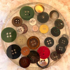May include: A clear, round resin coaster embedded with a variety of colorful buttons. The buttons are in shades of green, brown, black, red, and yellow. Some buttons have glitter accents.
