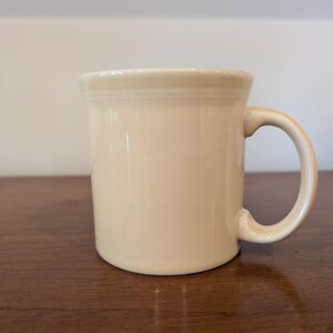 May include: A cream-colored ceramic mug with a rounded handle. The mug has a smooth, glossy finish and a simple, classic design. The mug is sitting on a wooden surface.