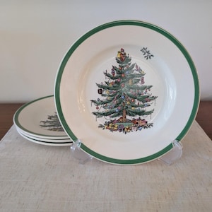 May include: Set of four white ceramic plates with a green border. Each plate features a detailed Christmas tree design in the center, decorated with ornaments and gifts. The plates are displayed on a neutral-colored linen surface.