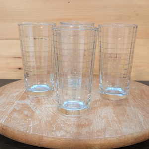 May include: A set of three clear glass tumblers, each featuring a textured grid pattern of horizontal and vertical lines etched into the exterior surface. The glasses have a cylindrical shape with a flat base and a smooth, transparent rim at the top.