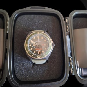 May include: A wristwatch with a black face, green bezel, and a gray strap, presented in a black protective case with foam padding. The watch features orange accents and is designed for diving.
