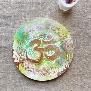Peut inclure: Une assiette ronde en bois avec un motif de mandala vert, brun et blanc et un symbole Om de couleur cuivre au centre.