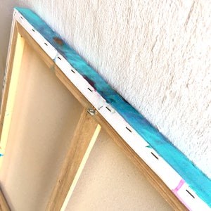 May include: Close-up of the back of a stretched canvas painting. The canvas is wrapped around a wooden frame and secured with staples. The painting features a blue, green, and pink abstract design.